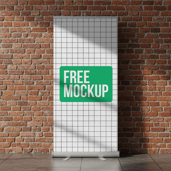 Brick Wall Roll Up Banner Free Mockup. A vertical roll-up banner stand with the words "Free Mockup" is placed against an exposed red brick wall, with sunlight casting shadows across the scene.