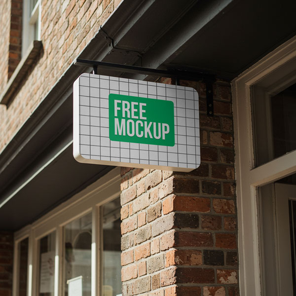 Hanging Storefront Sign Free Mockup. A horizontal hanging sign with rounded corners and a company logo is mounted on a black bracket against a red brick building facade, above a shop window.